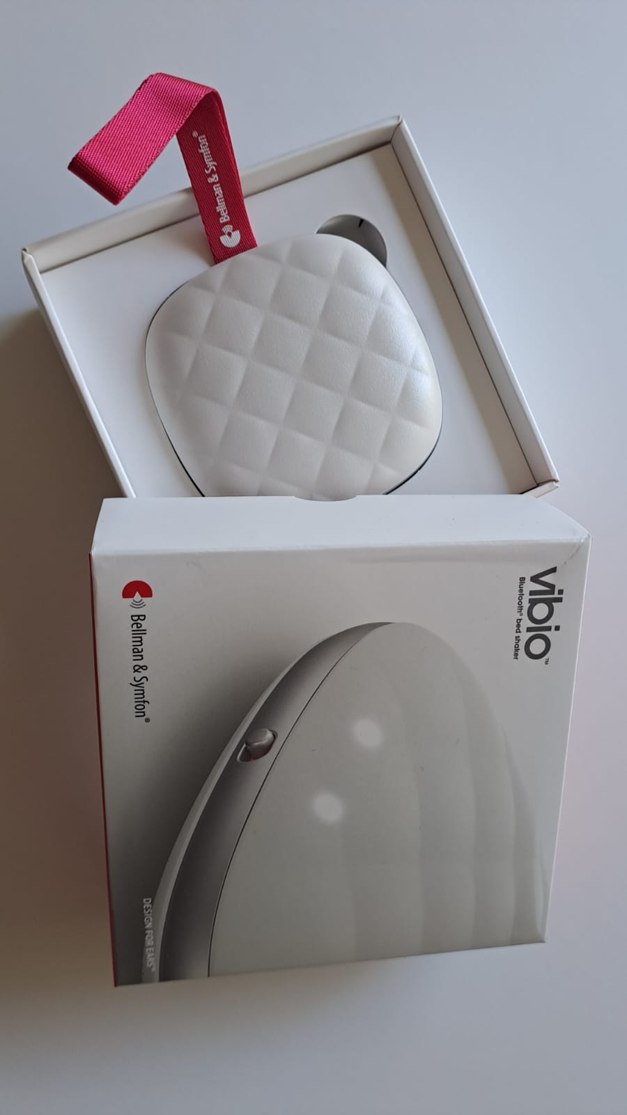 Otwarte pudełko poduszki wibracyjnej Vibio (Bluetooth bed shaker) firmy Bellman & Symfon. W środku biała, pikowana poduszka z czerwonym uchwytem z taśmy.