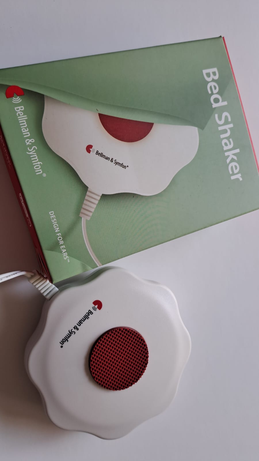 Wibrator pod poduszkę Bed Shaker firmy Bellman & Symfon – biała podkładka w kształcie kwiatu z czerwonym środkiem i przewodem, obok zielone opakowanie.