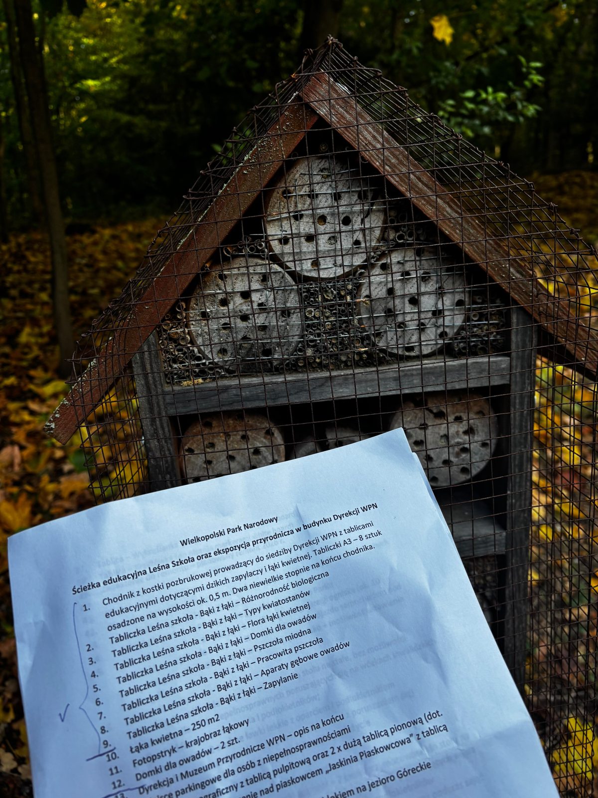 Wydruk listy elementów do inwentaryzacji „Ścieżka edukacyjna Leśna Szkoła oraz ekspozycja przyrodnicza w budynku Dyrekcji WPN” z zaznaczonymi pozycjami, trzymany przed domkiem dla owadów.