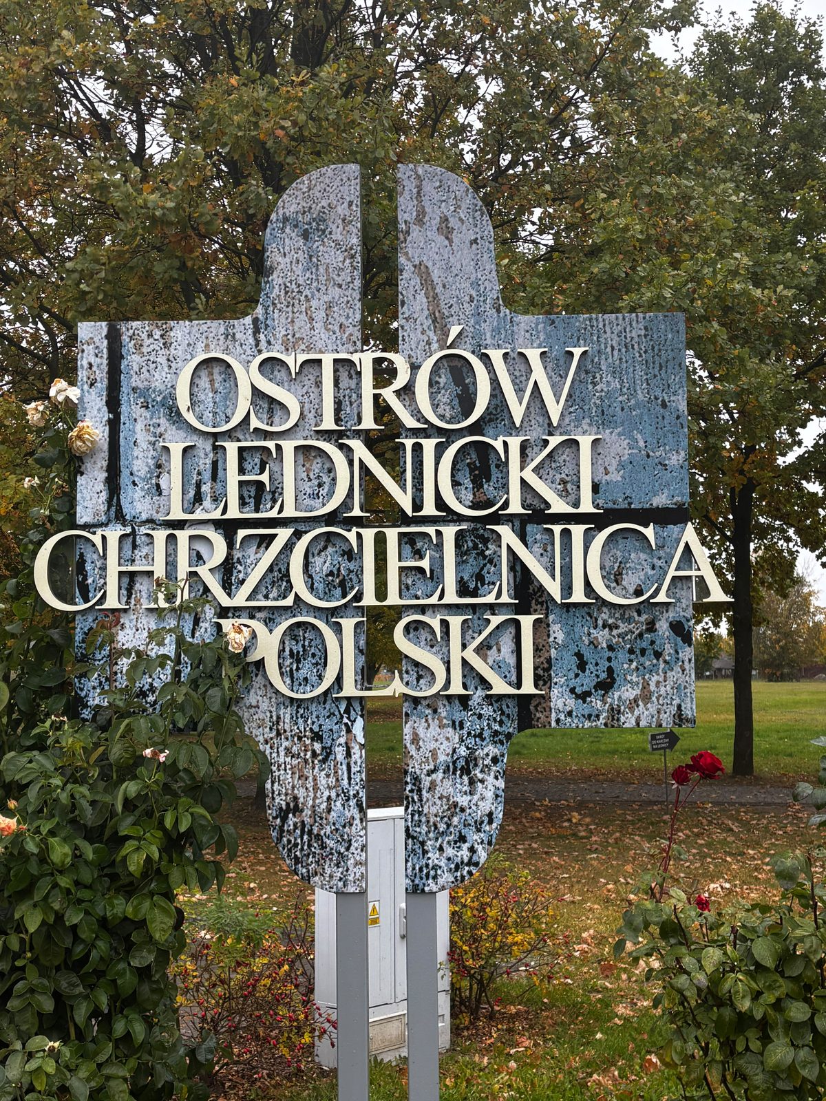 Znak w kształcie krzyża z napisem „Ostrów Lednicki Chrzcielnica Polski” wśród krzewów róż, w tle drzewa w jesiennych barwach.