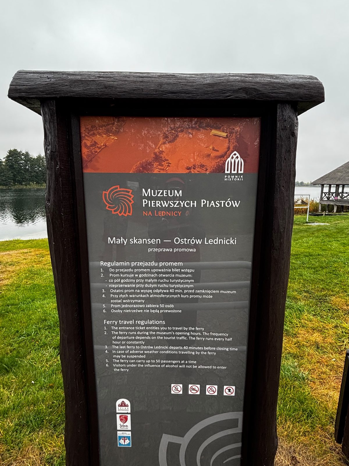 Tablica Muzeum Pierwszych Piastów na Lednicy „Mały skansen — Ostrów Lednicki, przeprawa promowa” z regulaminem przejazdu promem po polsku i angielsku; w tle jezioro i drewniana altana.