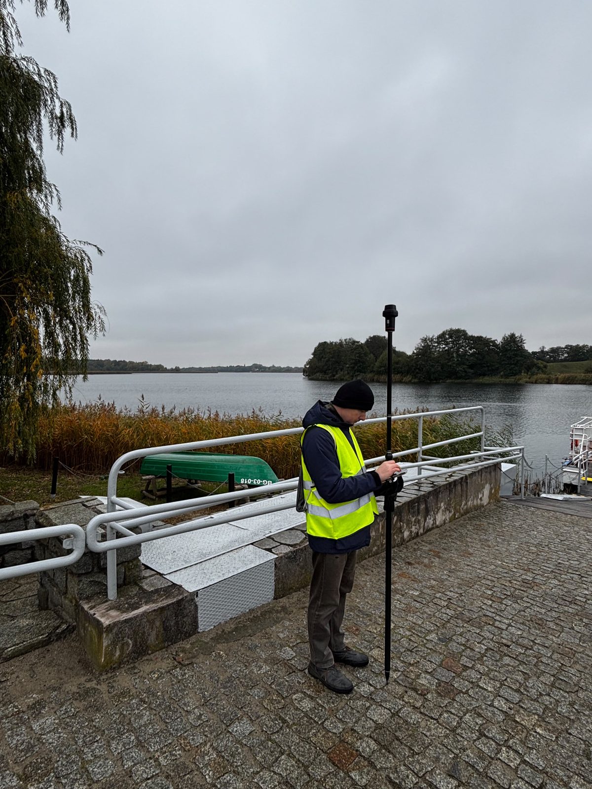 Osoba z zespołu w kamizelce odblaskowej wykonuje pomiar odbiornikiem GNSS na tyczce na brukowanym nabrzeżu przy przeprawie promowej; za nią pochylnia z poręczami, trzciny i jezioro.