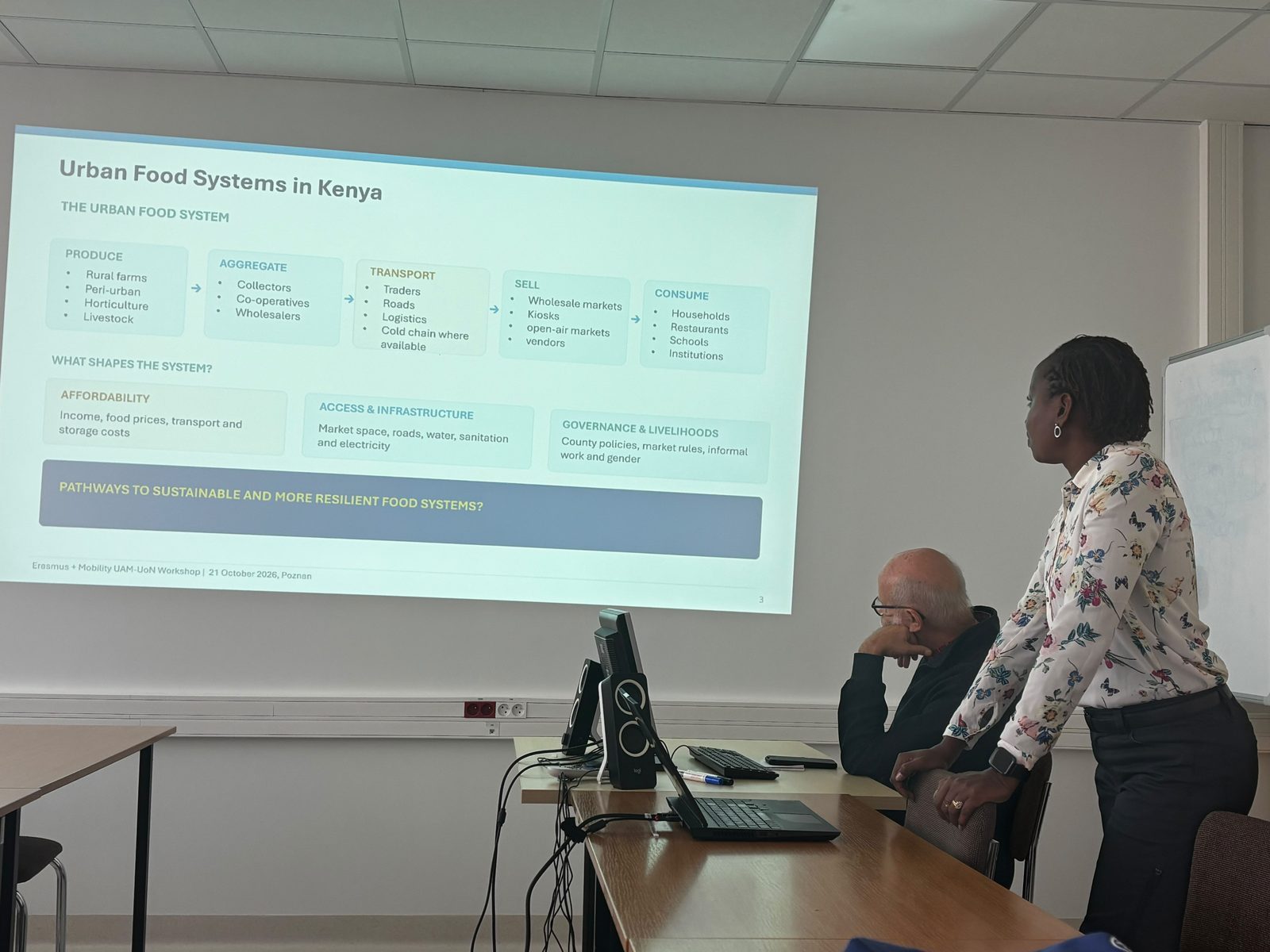 Osoba stojąca przy stole prezentuje slajd „Urban Food Systems in Kenya” z dopiskiem o warsztatach Erasmus+ UAM-UoN 21 października 2026 r. w Poznaniu; obok przy stole siedzi kolejna osoba.