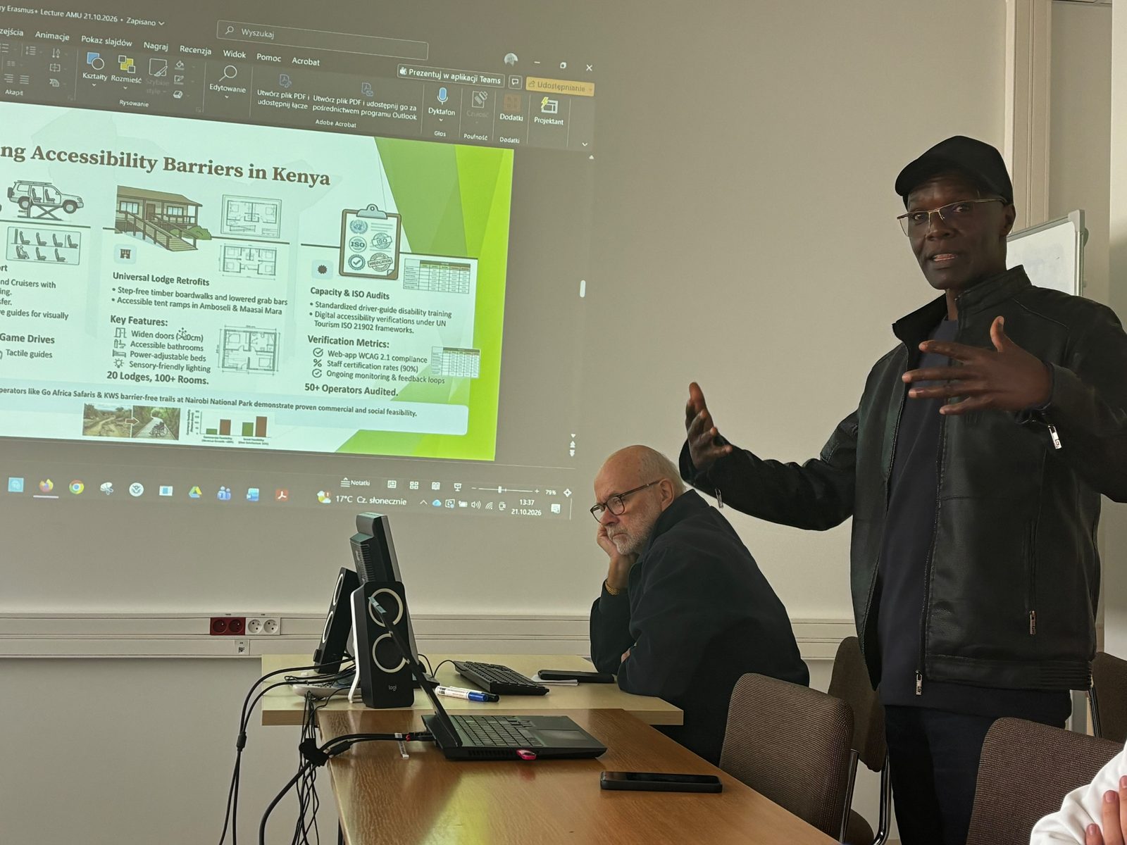 Kennedy Omoke z University of Nairobi podczas wystąpienia; w tle projekcja slajdu o barierach dostępności w turystyce w Kenii, przy stole siedzi słuchająca osoba.