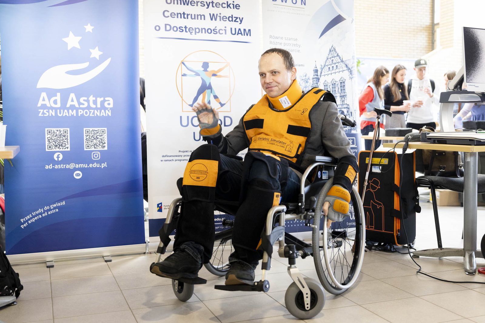 Mężczyzna w kostiumie geriatrycznym, z kołnierzem ortopedycznym i nakładkami na nogach, siedzi na wózku i macha ręką; w tle roll-upy Ad Astra ZSN UAM, UCWoD UAM i Biura Wsparcia Osób z Niepełnosprawnościami.